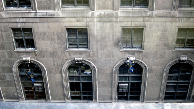 Window-washers, viewed from above, spider down the facade of an old stone building.
