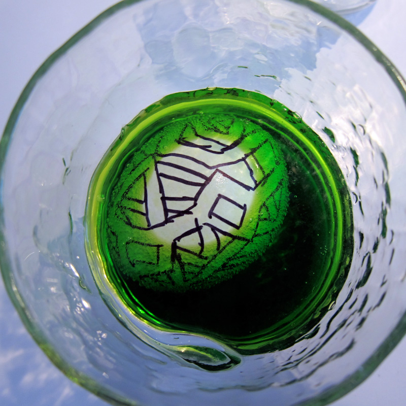 An Easter egg with an Escher-like drawing on it soaking in a glass of green egg dye.